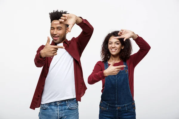 Feliz joven pareja afroamericana mirando a través de un marco de dedo y sonriendo mientras está de pie aislado en blanco . —  Fotos de Stock
