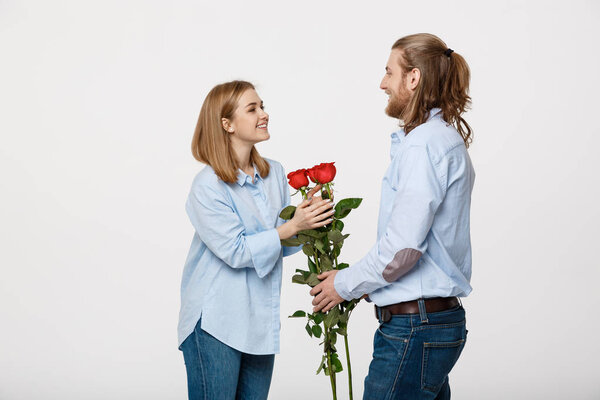 Portrait of handsome elegant guy is surprising his beautiful girlfriend with red roses and smiling over white isolated background.