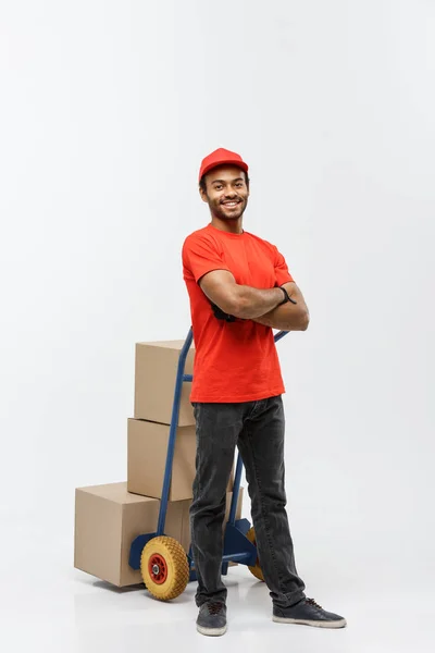 Concept de livraison - Portrait de beau livreur afro-américain ou messager poussant camion à main avec pile de boîtes. Isolé sur fond de studio Grey. Espace de copie . — Photo