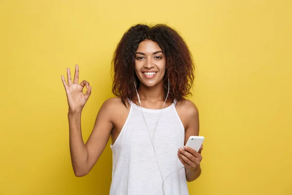 Lifestyle Concept - Retrato de la hermosa mujer afroamericana alegre escuchando música en el teléfono móvil y mostrar signo ok con los dedos. Fondo amarillo pastel estudio. Copiar espacio . — Foto de Stock