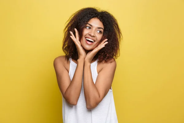 Glimlachend mooie jonge African American vrouw in het witte T-shirt poseren met op de kin handen. Studio opname op gele achtergrond. Kopiëren van ruimte. — Stockfoto