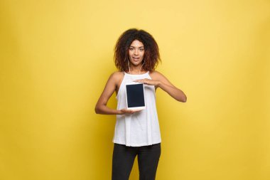 Yaşam tarzı kavramı - güzel Afrikalı-Amerikalı kadın bir şey elektronik tablet üzerinde sunulması neşeli portre. Sarı pastel studio arka plan. Kopya alanı.