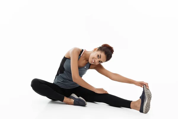 Deporte, entrenamiento, estilo de vida y concepto de fitness: retrato de la hermosa mujer afroamericana feliz estirando la pierna mientras está sentada. Aislado sobre fondo blanco estudio . —  Fotos de Stock