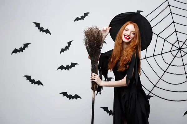 Halloween heks Concept - portret van mooie jonge heks met bezem over grijs muur met vleermuis en spider web-achtergrond. — Stockfoto