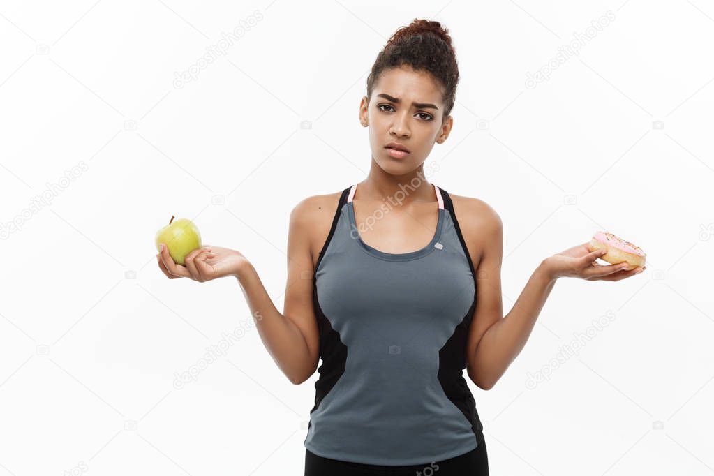 Healthy and diet concept - Beautiful sporty African American make a decision between donut and green apple. Isolated on white background.