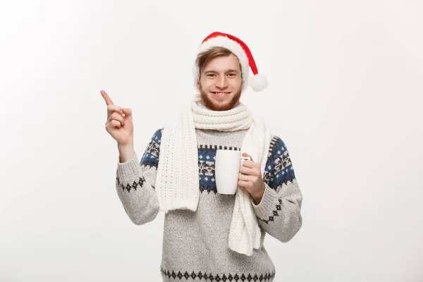 Weihnachtskonzept - junger Mann mit Bart, Pullover und Weihnachtsmütze, der eine heiße Kaffeetasse hält und die Hand auf die weiße Seite zeigt, mit Kopierraum. — Stockfoto