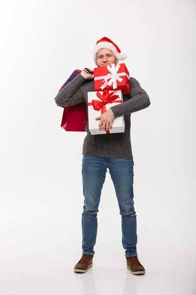 Christmas Concept - young handsome man with beard holding heavy presents and shopping bags with exhausted facial expression on white background. — Stock Photo, Image