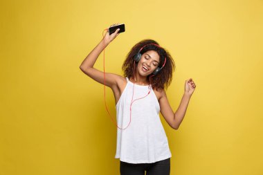 Yaşam Tarzı Konsepti - Cep telefonundan müzik dinleyen güzel Afro-Amerikan kadın portresi. Sarı pastel stüdyo arka planı. Boşluğu Kopyala