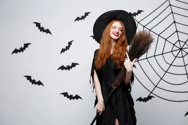 Halloween čarodějnice koncept - portrét krásné mladé čarodějnice s koště nad šedou zeď s bat a spider webové pozadí. — Stock fotografie