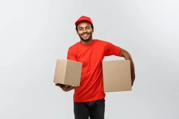 Conceito de entrega - Retrato de homem de entrega afro-americano feliz em pano vermelho segurando um pacote de caixa. Isolado no estúdio Grey Background. Espaço de cópia . — Fotografia de Stock