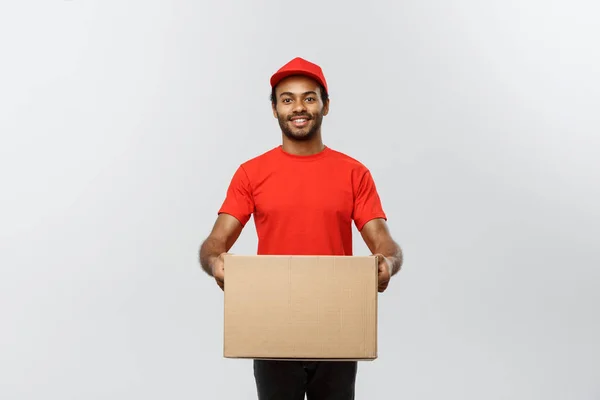 Conceito de entrega - Retrato de homem de entrega afro-americano feliz em pano vermelho segurando um pacote de caixa. Isolado no estúdio Grey Background. Espaço de cópia . — Fotografia de Stock