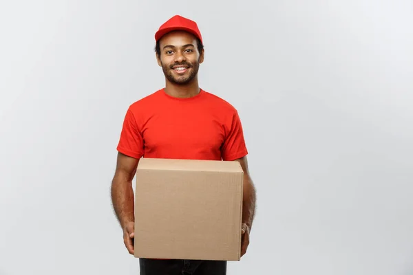 Conceito de entrega - Retrato de homem de entrega afro-americano feliz em pano vermelho segurando um pacote de caixa. Isolado no estúdio Grey Background. Espaço de cópia . — Fotografia de Stock