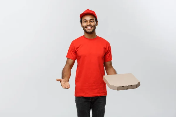 Concetto di consegna Ritratto dell'uomo di consegna afroamericano felice che punta la mano per presentare un pacchetto di scatole di pizza. Isolato su sfondo grigio studio. Copia spazio . — Foto Stock