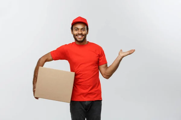 Concetto di consegna Ritratto dell'uomo di consegna afroamericano felice che punta la mano per presentare un pacchetto scatola. Isolato su sfondo grigio studio. Copia spazio . — Foto Stock