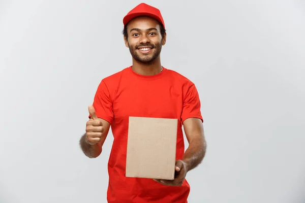 Concepto de entrega - Retrato del repartidor afroamericano feliz sosteniendo un paquete de caja y mostrando golpes. Aislado en Grey studio Background. Copiar espacio . — Foto de Stock