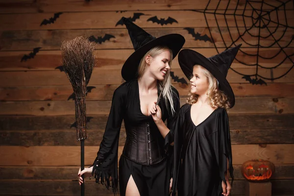 Halloween-Konzept - fröhliche Mutter und ihre Tochter in Hexenkostümen, die mit geschwungenen Kürbissen über Fledermäusen und Spinnennetz auf hölzernem Studiohintergrund Halloween feiern. — Stockfoto