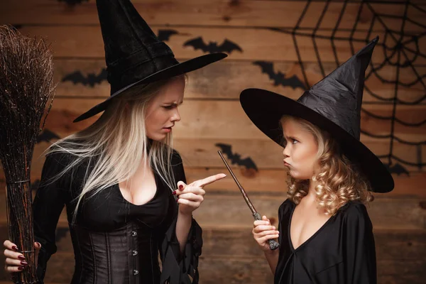 Concepto de Halloween - madre bruja estresante enseñando a su hija en trajes de bruja celebrando Halloween sobre murciélagos y tela de araña en fondo de estudio de madera . — Foto de Stock