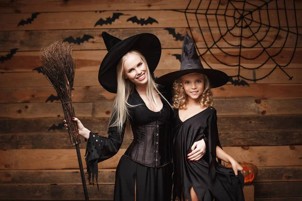 Concepto de Halloween - madre alegre y su hija en trajes de bruja celebrando Halloween posando con calabazas curvas sobre murciélagos y tela de araña sobre fondo de estudio de madera . —  Fotos de Stock