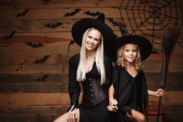 Concepto de Halloween - madre alegre y su hija en trajes de bruja celebrando Halloween posando con calabazas curvas sobre murciélagos y tela de araña sobre fondo de estudio de madera . — Foto de Stock