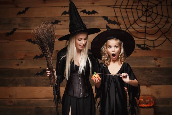 Concepto de Halloween - hermosa madre caucásica y su hija en trajes de bruja disfrutan usando magia con varita mágica para tarro de calabaza de Halloween sobre murciélagos y tela de araña en fondo de estudio de madera . — Foto de Stock