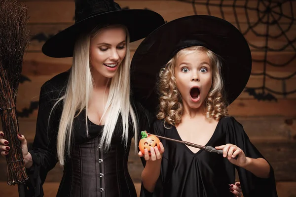 Halloween-Konzept - schöne kaukasische Mutter und ihre Tochter in Hexenkostümen genießen es, mit Zauberstab zu Halloween Kürbiskrüge über Fledermäuse und Spinnennetz auf hölzernem Studiohintergrund zu zaubern. — Stockfoto