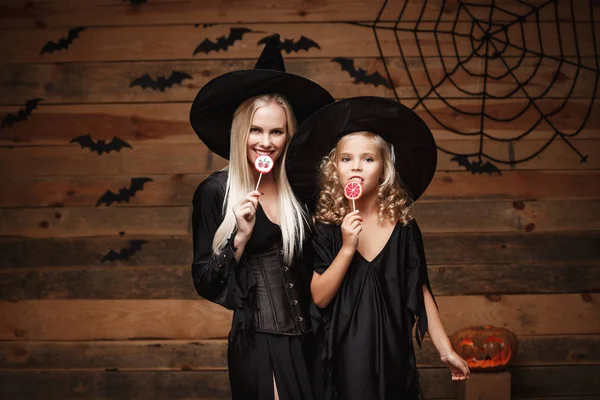 Halloween Concept - bella madre caucasica e sua figlia in costumi da strega che festeggiano Halloween con caramelle di Halloween e dolci su pipistrelli e ragnatela su sfondo in legno . — Foto Stock