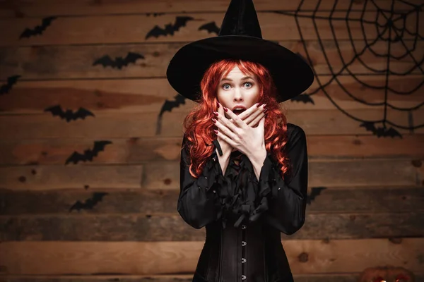 Halloween heks concept - Happy Halloween rood haar heks bedrijf poseren met gezicht geschokt over oude houten studio achtergrond. — Stockfoto