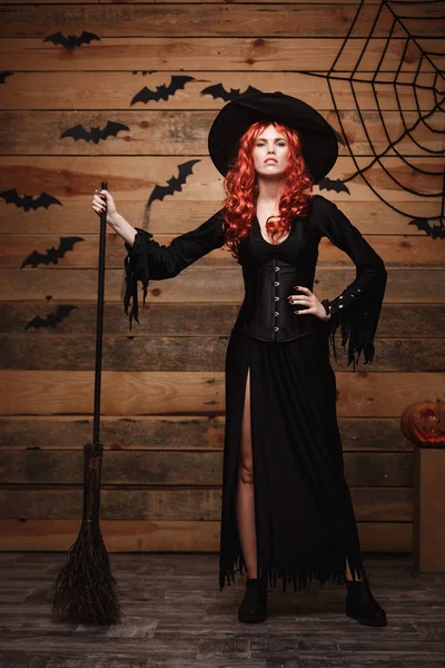 Halloween heks concept - Full-length Happy Halloween rode haren Witch holding poseren met magische bezemsteel op oude houten studio achtergrond. — Stockfoto