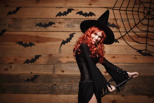 Conceito de bruxa de Halloween - Feliz Halloween cabelo vermelho Bruxa segurando vassoura mágica voando gesto sobre velho estúdio de madeira fundo . — Fotografia de Stock