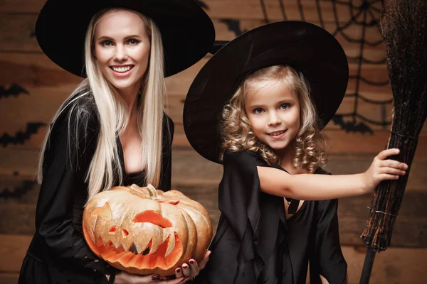 Concepto de Halloween - Primer plano hermosa madre caucásica y su hija en trajes de bruja celebrando Halloween posando con calabazas curvas sobre los murciélagos y tela de araña sobre fondo de estudio de madera . — Foto de Stock