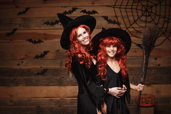 Mooie Kaukasische moeder en haar dochter met lange rode haren in heks kostuums vieren Halloween poseren met over vleermuizen en spinnenweb op houten studio achtergrond. — Stockfoto