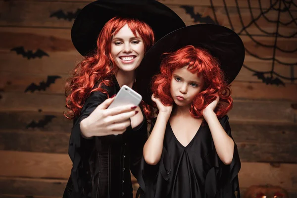 Koncepcja Halloween - piękne kaukaski matka i jej córka z długimi rudymi włosami w stroje czarownic biorąc selfie z smartphone gotowy do odprawiania Halloween. — Zdjęcie stockowe