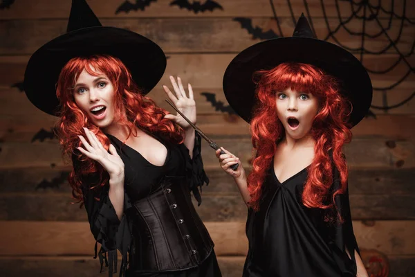 Mooie Kaukasische moeder en haar dochter met lange rode haren in heks kostuums en magische toverstaf vieren Halloween poseren met over vleermuizen en spinnenweb op houten studio achtergrond. — Stockfoto