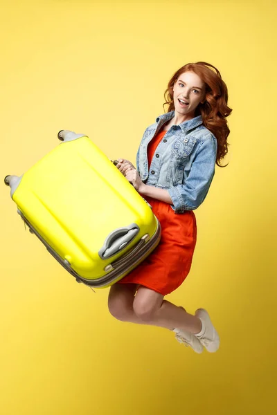 Travel and Lifestyle Concept: Retrato de uma bela mulher de moda de cabelo vermelho pulando e segurando uma mala verde brilhante isolada sobre fundo amarelo brilhante . — Fotografia de Stock