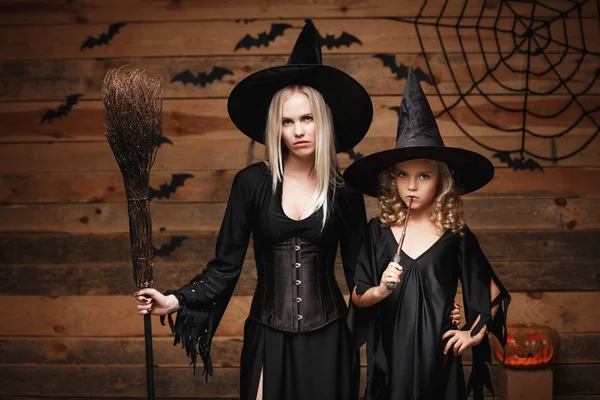 Concepto de Halloween - madre alegre y su hija en trajes de bruja celebrando Halloween posando con calabazas curvas sobre murciélagos y tela de araña sobre fondo de estudio de madera . — Foto de Stock