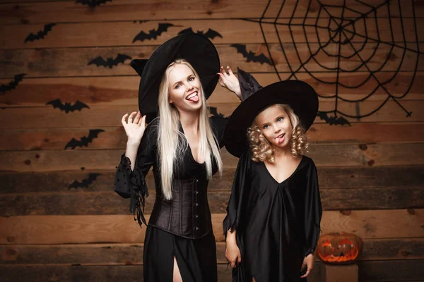 Concepto de Halloween - madre alegre y su hija en trajes de bruja celebrando Halloween posando con calabazas curvas sobre murciélagos y tela de araña sobre fondo de estudio de madera . — Foto de Stock