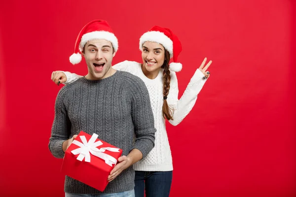 Рождественская концепция - молодая женщина закрывает глаза мужчин рукой и делает сюрприз большой подарок. Изолированный на красном фоне — стоковое фото