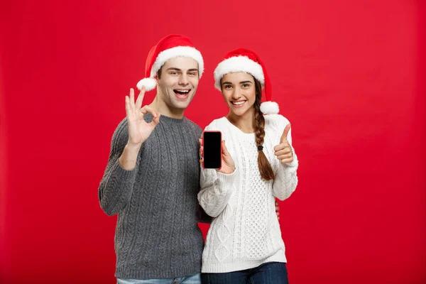 Conceito de Natal - Jovem casal feliz em camisolas de Natal mostrando gesto ok com telefone celular — Fotografia de Stock