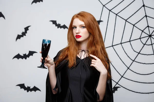 Halloween witch concept - Happy Halloween Witch holding glass of bloody red wine over dark grey studio background with bat and spider web.