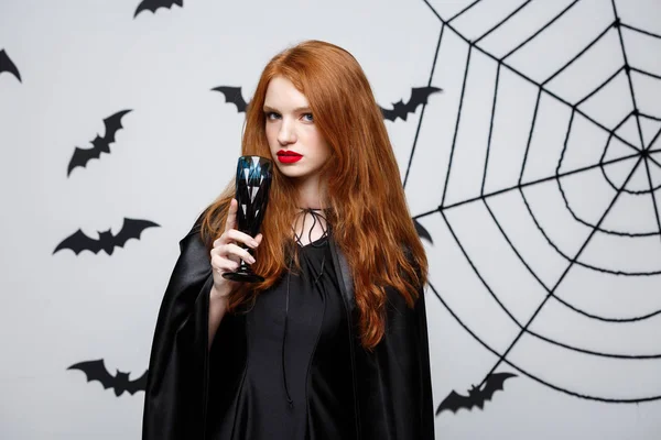 Απόκριες μάγισσα έννοια - ευτυχής Απόκριες μάγισσα κρατώντας το ποτήρι αιματηρή κόκκινο κρασί πάνω από φόντο σκούρο γκρι στούντιο με ρόπαλο και spider web. — Φωτογραφία Αρχείου