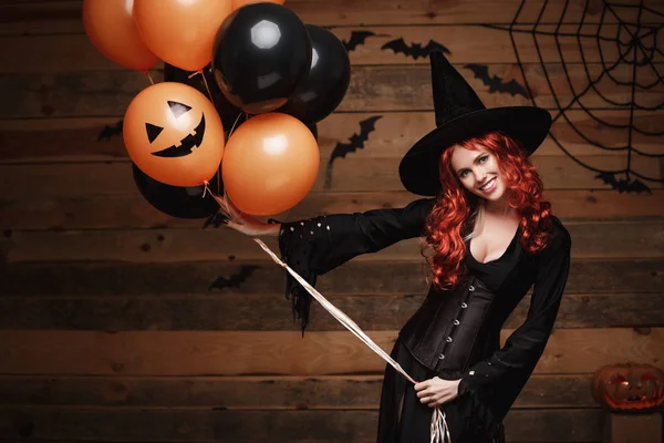 Halloween heks Concept - mooie Kaukasische vrouw in heks kostuums vieren Halloween poseren met poseren met oranje en zwarte ballon over vleermuizen en spinnenweb op houten studio achtergrond. — Stockfoto