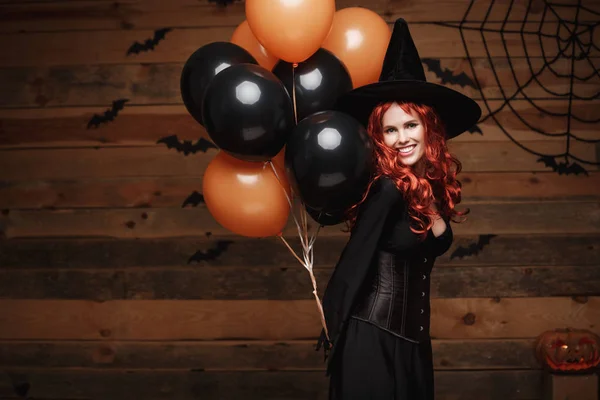 Halloween heks Concept - mooie Kaukasische vrouw in heks kostuums vieren Halloween poseren met poseren met oranje en zwarte ballon over vleermuizen en spinnenweb op houten studio achtergrond. — Stockfoto
