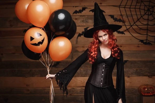 Halloween heks Concept - mooie Kaukasische vrouw in heks kostuums vieren Halloween poseren met poseren met oranje en zwarte ballon over vleermuizen en spinnenweb op houten studio achtergrond. — Stockfoto