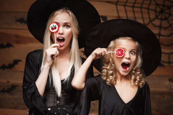 Halloween concept - schöne kaukasische Mutter und ihre Tochter in Hexenkostümen feiern halloween mit halloween candy und sweet über Fledermäusen und Spinnennetz auf hölzernem Studiohintergrund. — Stockfoto