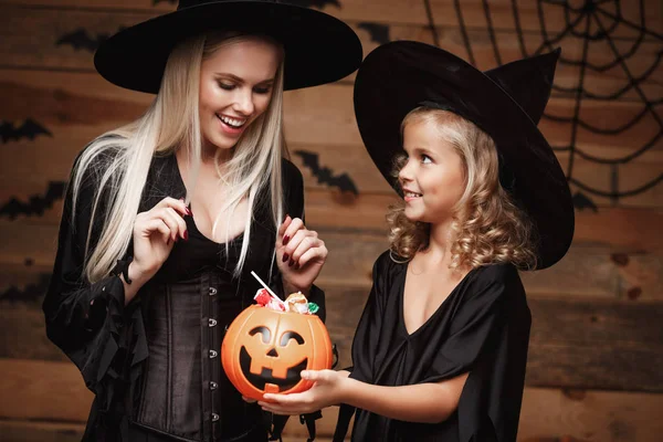 Halloween Concept - bella madre caucasica e sua figlia in costumi da strega che festeggiano Halloween con la condivisione di caramelle di Halloween e dolci su pipistrelli e ragnatela su sfondo in legno studio . — Foto Stock