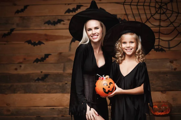 Halloween Concept - hermosa madre caucásica y su hija en trajes de bruja celebrando Halloween con el intercambio de dulces de Halloween y dulce sobre los murciélagos y la tela de araña en el fondo del estudio de madera . — Foto de Stock