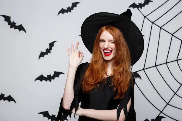 Halloween boszorkány koncepció - happy halloween gyömbér haj boszorkány tartja az ok jele, ujjaival, pózol, mint szürke háttér. — Stock Fotó