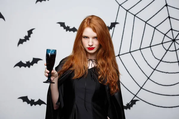 Halloween heks concept - Happy Halloween heks houdt glas bloedige rode wijn over donker grijs studio achtergrond met vleermuis en spider web. — Stockfoto