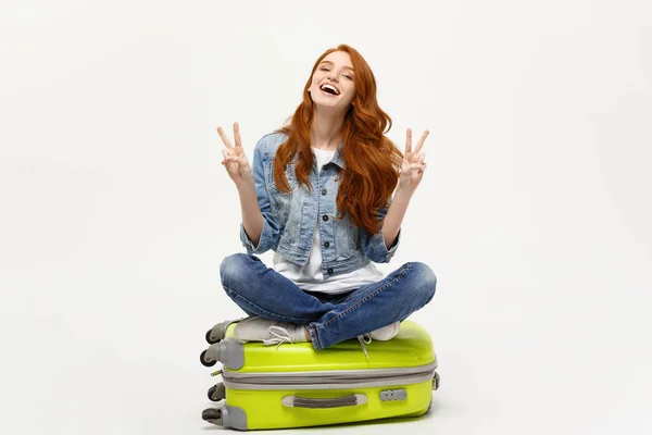 Conceito de viagem: jovem mulher branca sorridente sentada na mala mostrando dois dedos. Isolado sobre fundo branco . — Fotografia de Stock