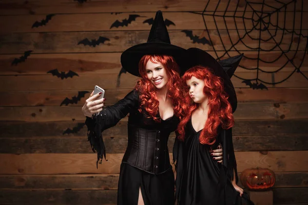 Концепция Хэллоуина - красивая кавказская мать и ее дочь с длинными рыжими волосами в костюмах ведьм делают селфи со смартфоном, готовым к празднованию Хэллоуина . — стоковое фото
